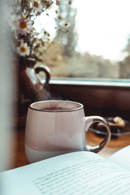 A steaming mug of tea by a sunlit window, with daisies and an open book.