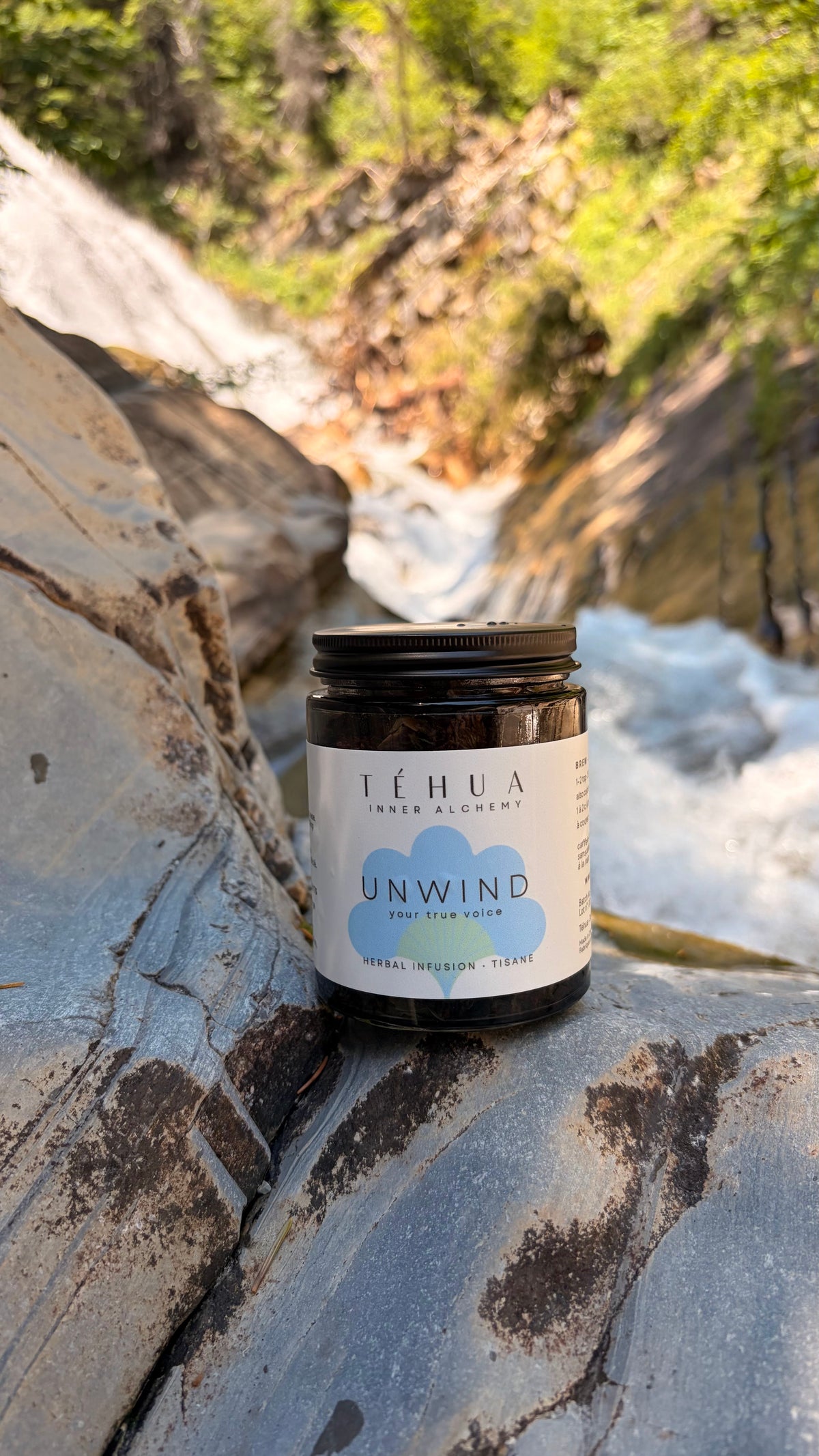 A TÉHUA Inner Alchemy UNWIND herbal tea jar on a large grey rock outdoors, with a blurred waterfall and forest behind.