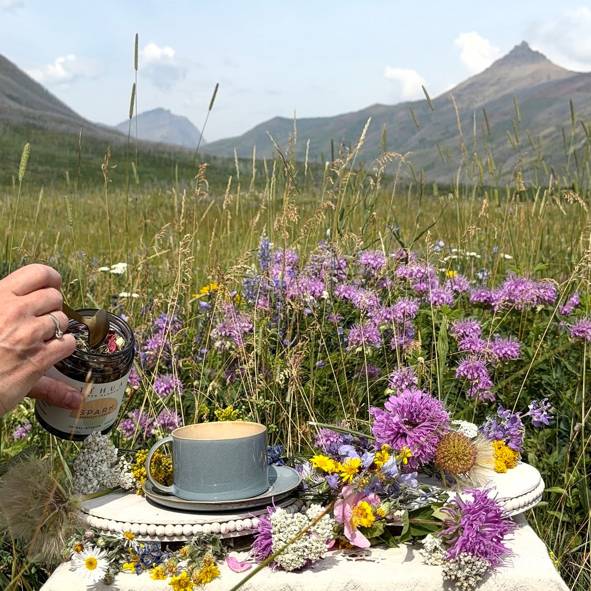 A hand pouring loose-leaf TÉHUA SPARK herbal tea from its jar into a blue-grey teacup, on a lace-covered table among wildflowers in a mountain meadow.