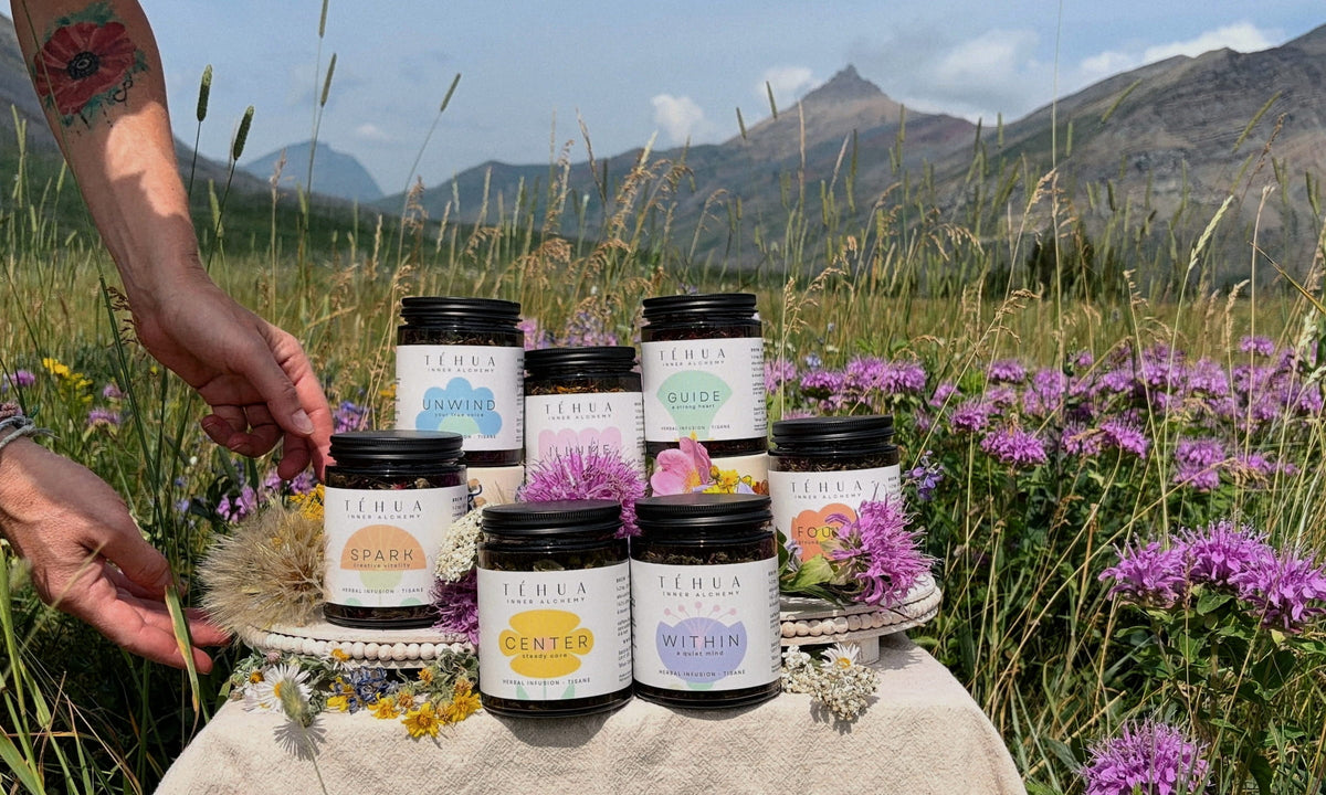 The seven TÉHUA herbal tea blends on a cloth-covered table in a mountain wildflower meadow, with a hand reaching in from the left.